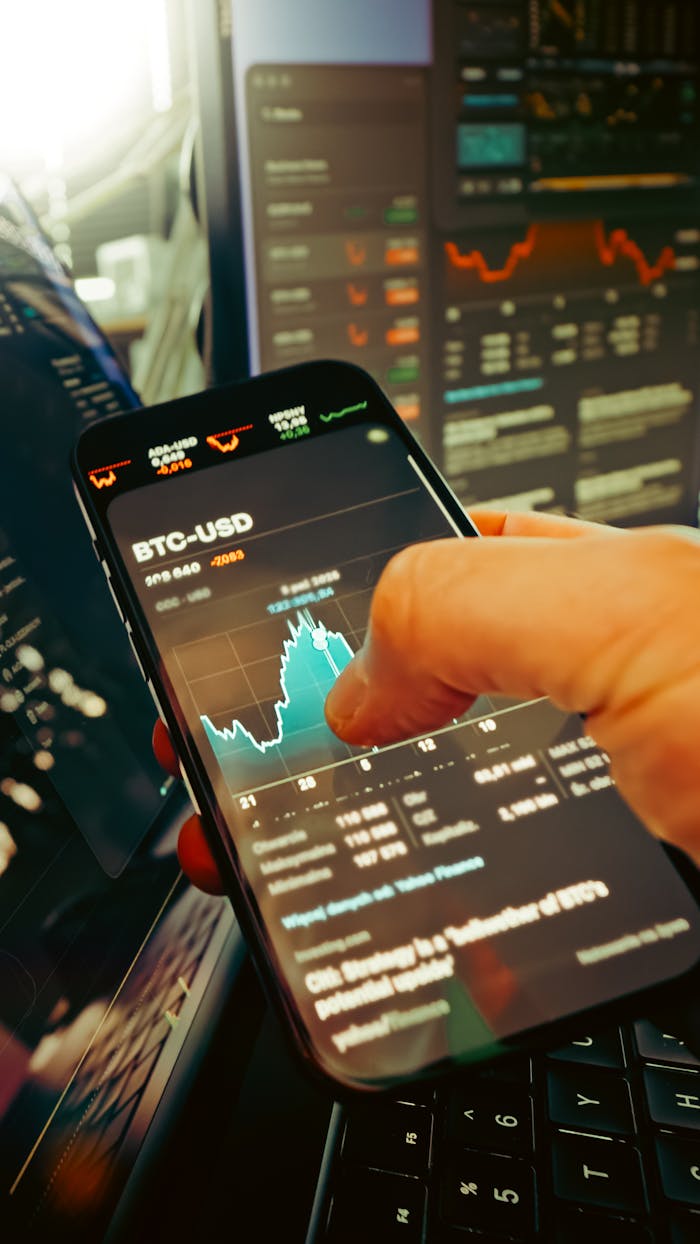 services-03 A hand holds a smartphone displaying BTC-USD chart amidst digital trading screens, highlighting crypto finance.