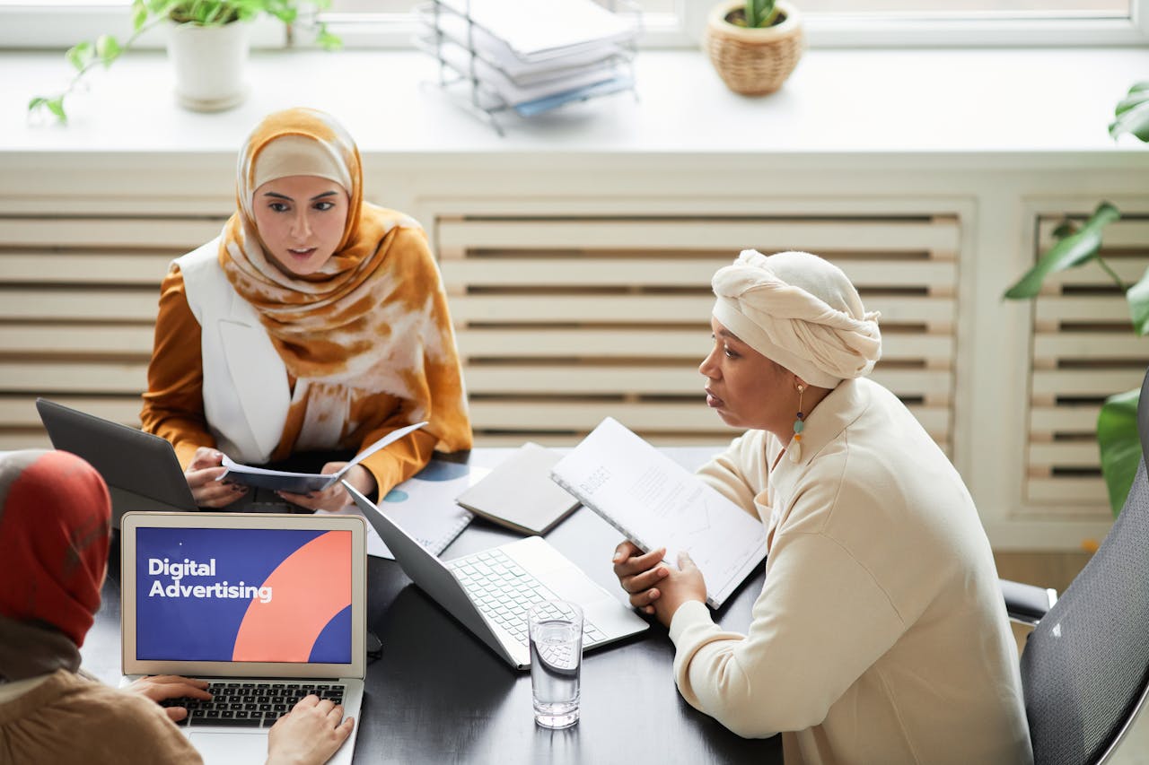 our-experience Three women in hijabs collaborating on digital advertising strategies around a table with laptops.